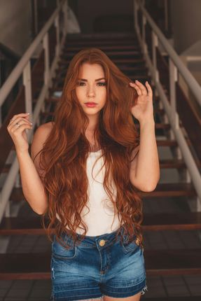 Woman with long red hair in denim shorts and white top, posing in front of a wooden staircase.