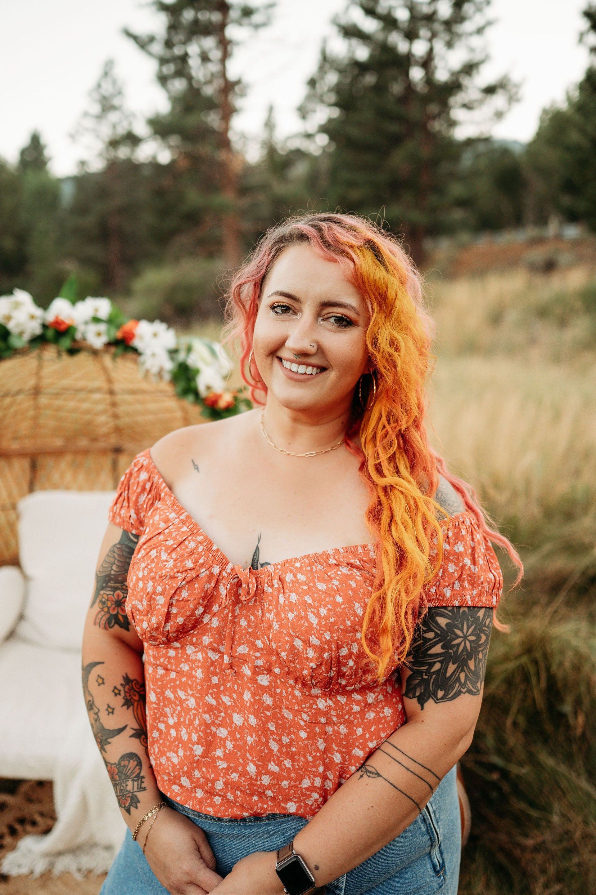 Woman with orange hair smiles outdoors; wearing a floral top and jeans, tattoos on arms.