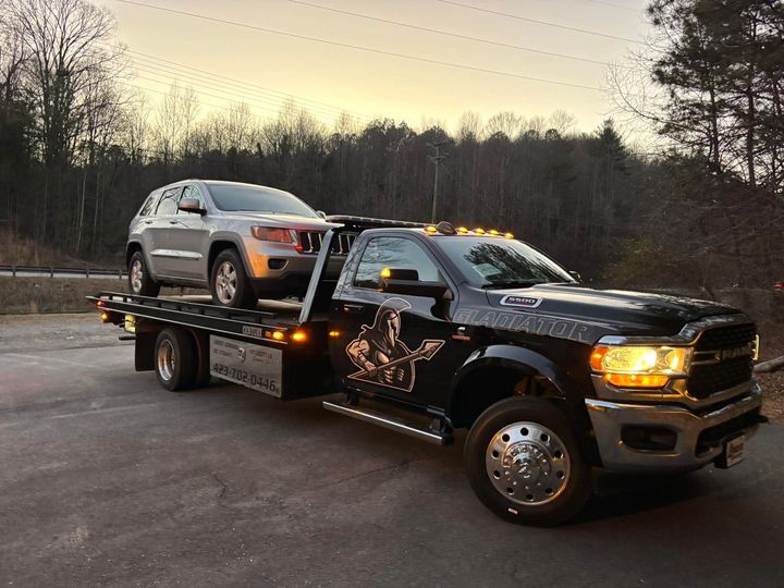 Black flatbed tow truck carrying a silver SUV on a roadside at dusk