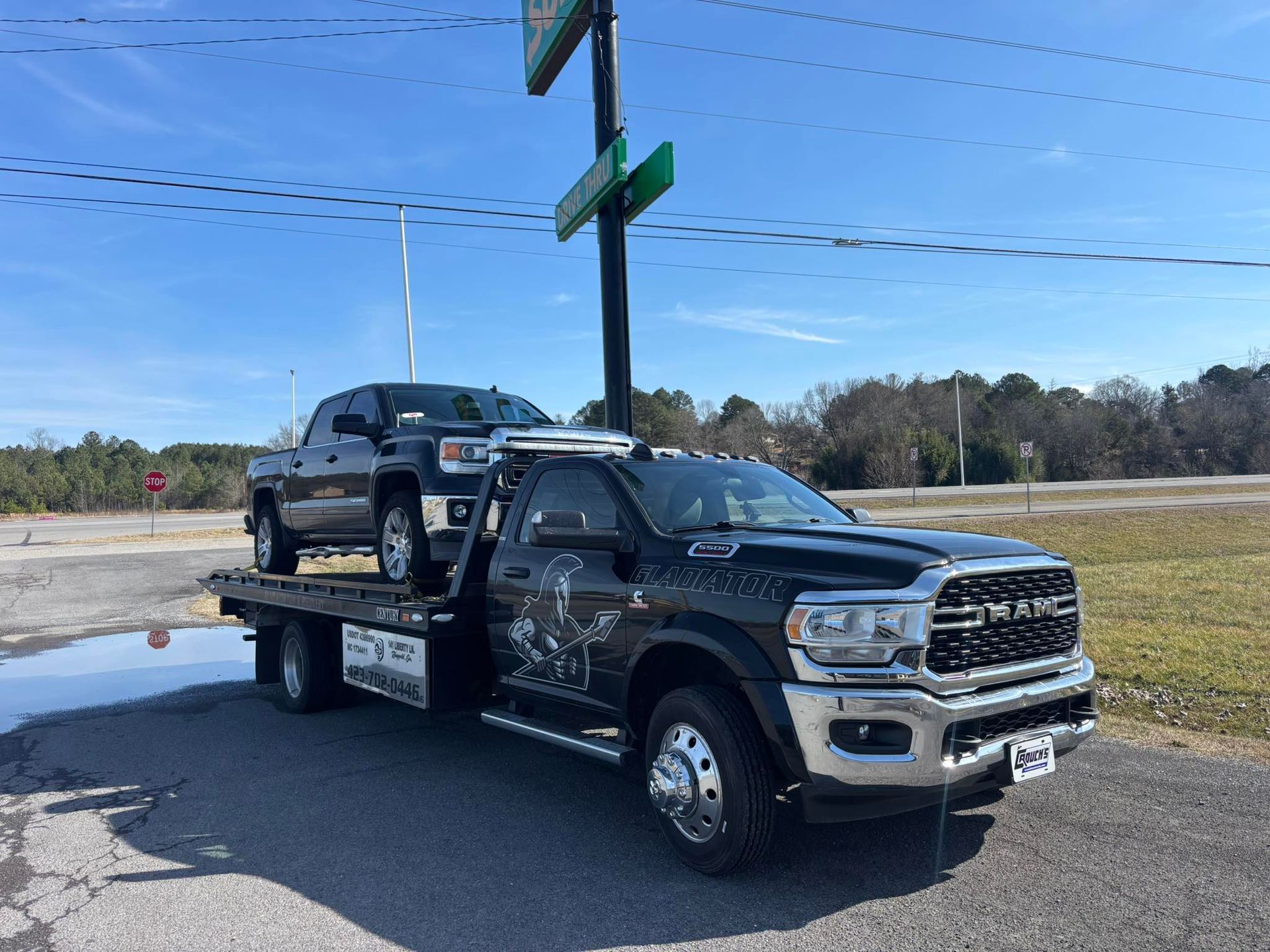 Black flatbed tow truck carrying a black pickup on a roadside under blue sky.