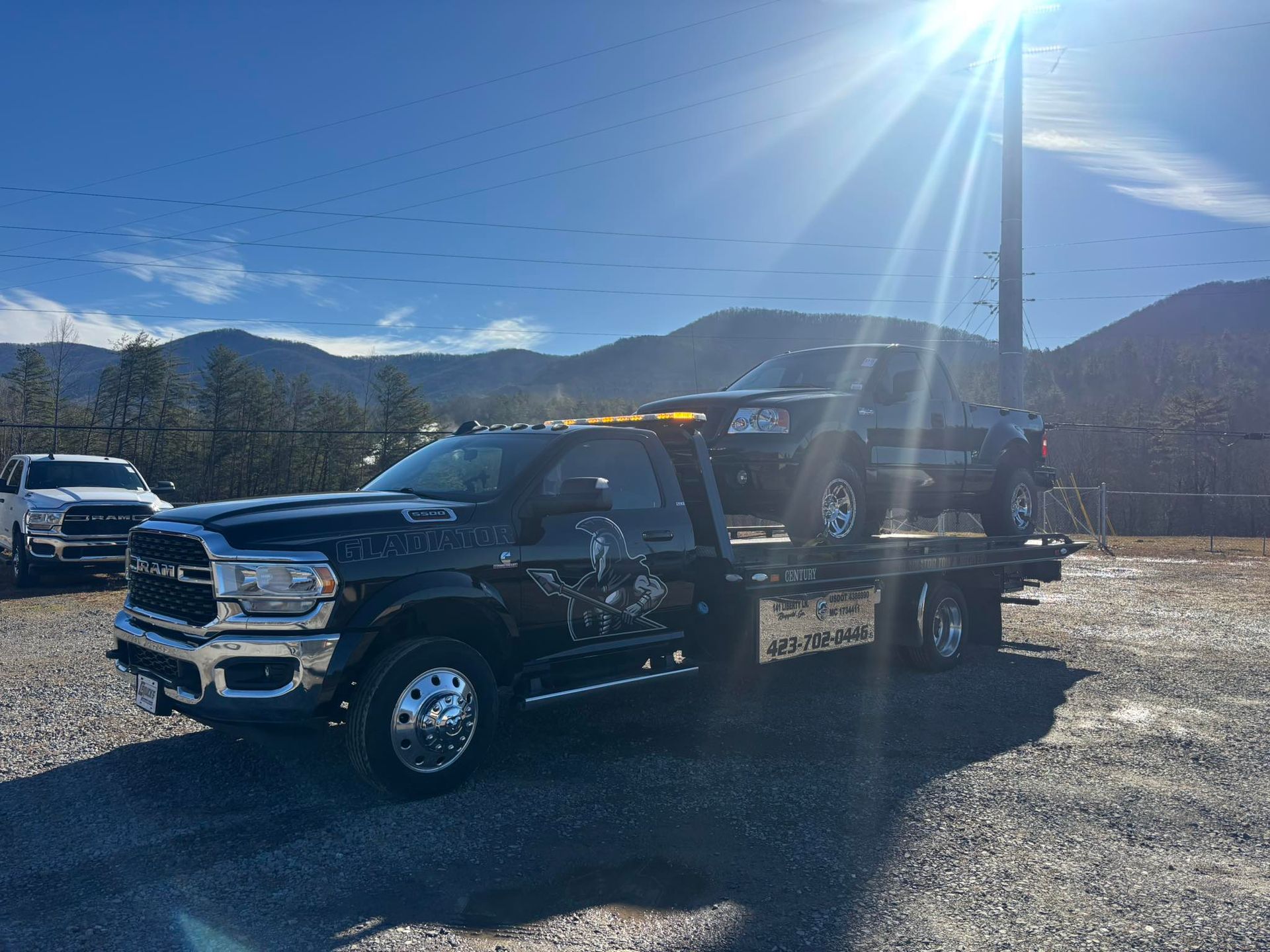 Black pickup truck towing a flatbed trailer on a gravel lot with mountains and bright sun in the background