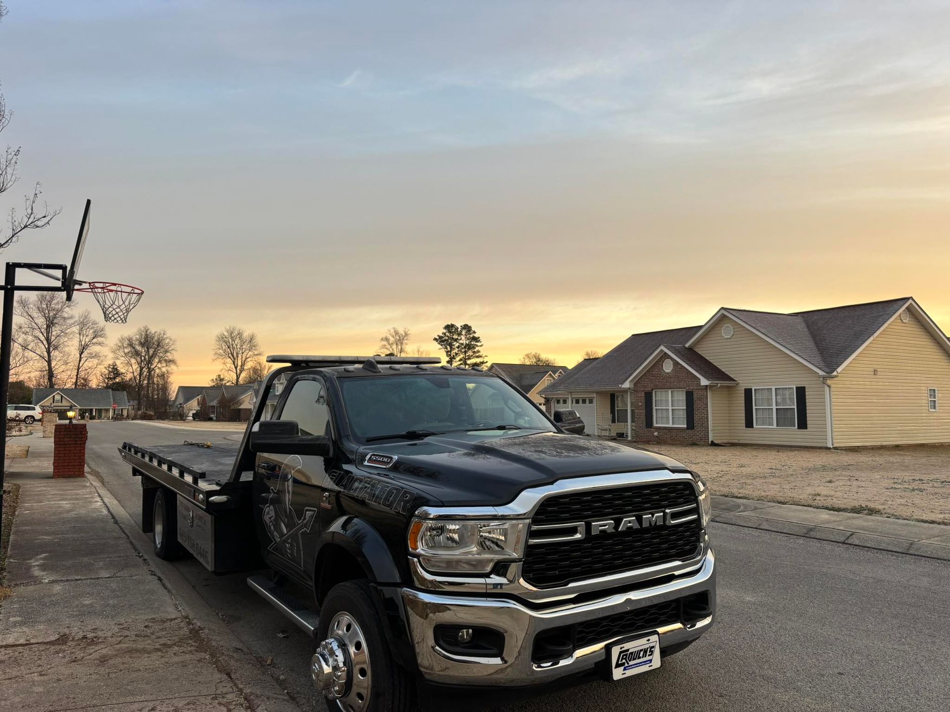 Black flatbed tow truck parked on a suburban street at sunset, with houses in the background