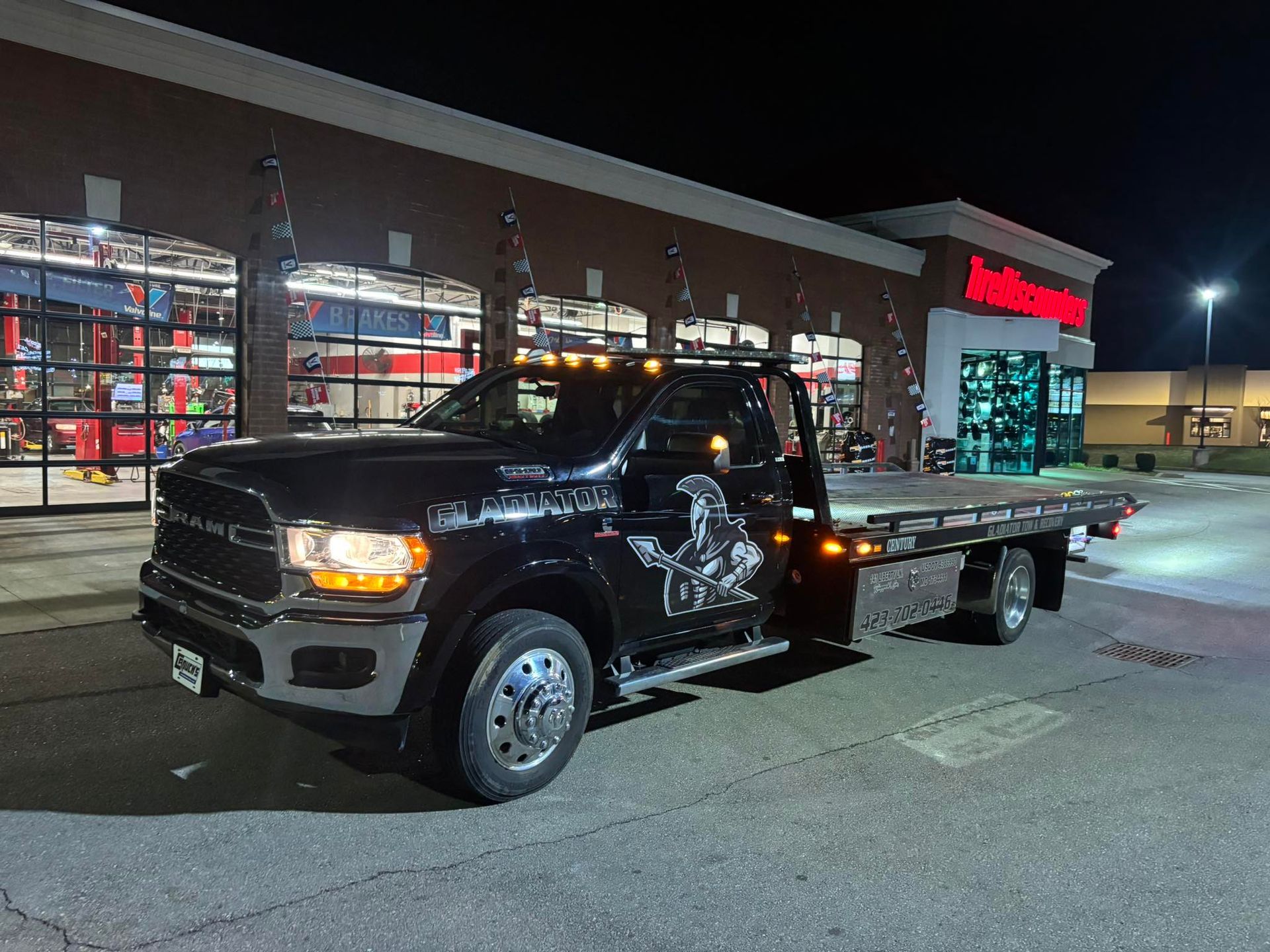 Black flatbed tow truck parked outside a store at night, headlights on.