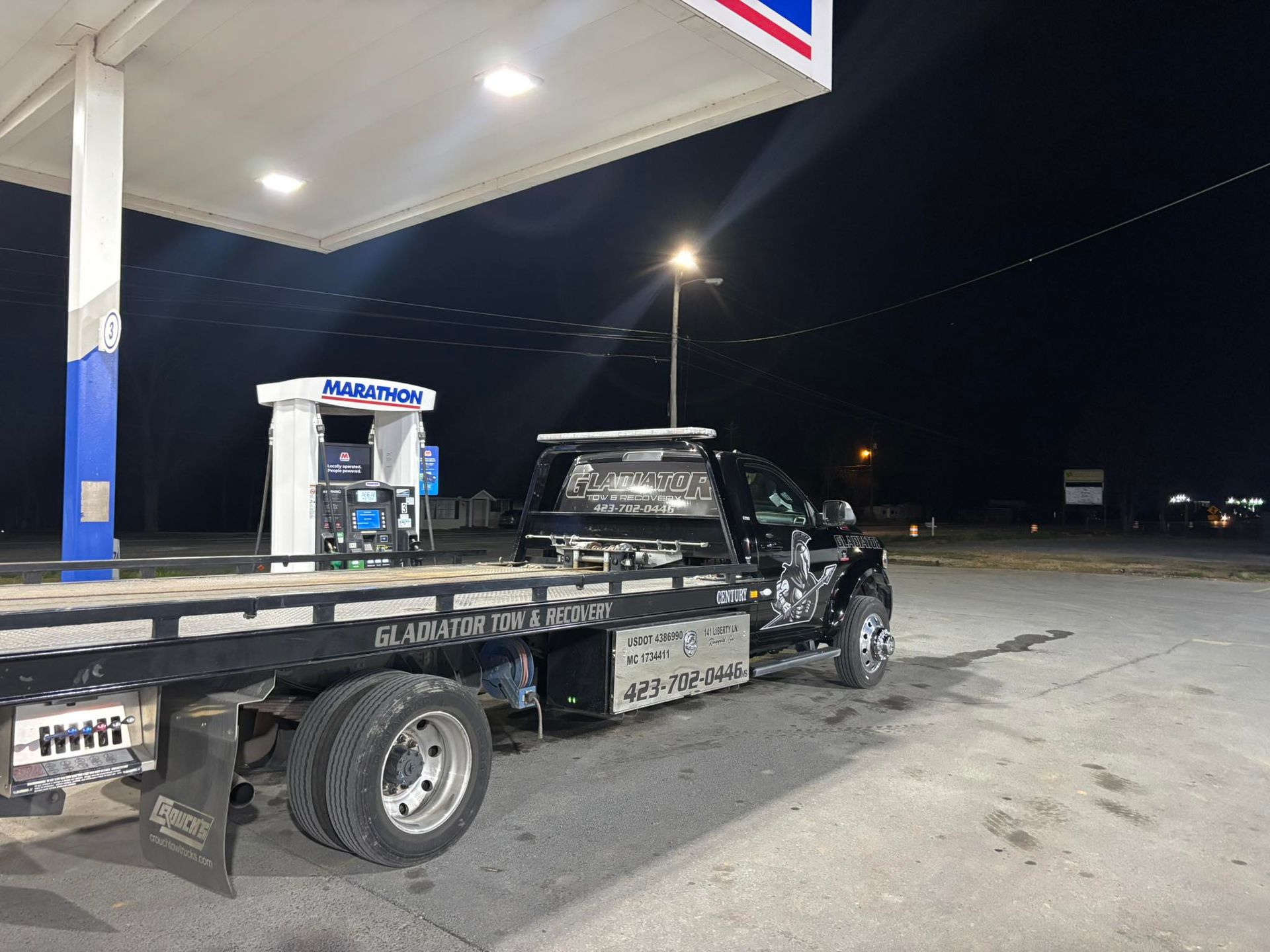 Tow truck at a gas station at night, parked beside the fuel pumps under bright canopy lights.
