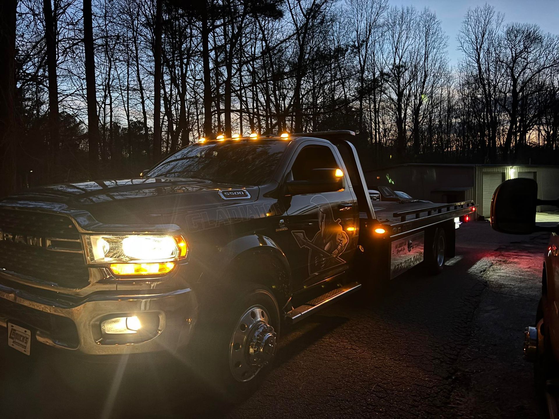 Black pickup truck with headlights on at dusk, parked on a road beside a dark car and bare trees.
