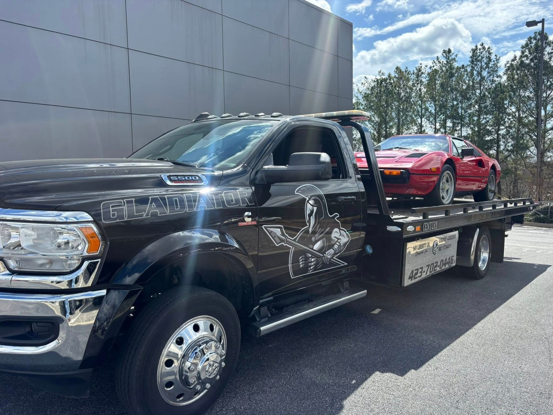 Black flatbed tow truck carrying a red sports car on a sunny road, beside a large building