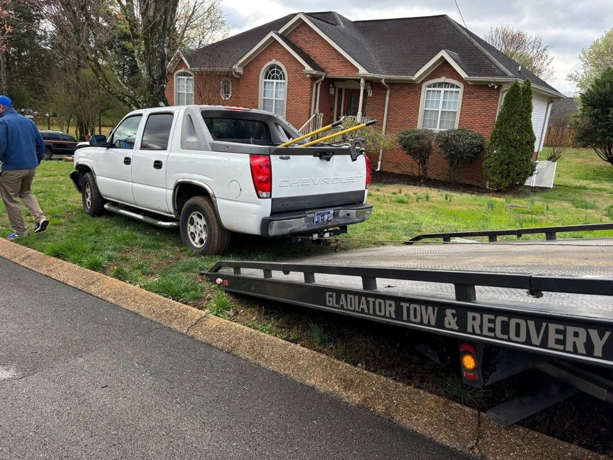 White pickup loaded on a tow truck in front of a brick house on a wet suburban street