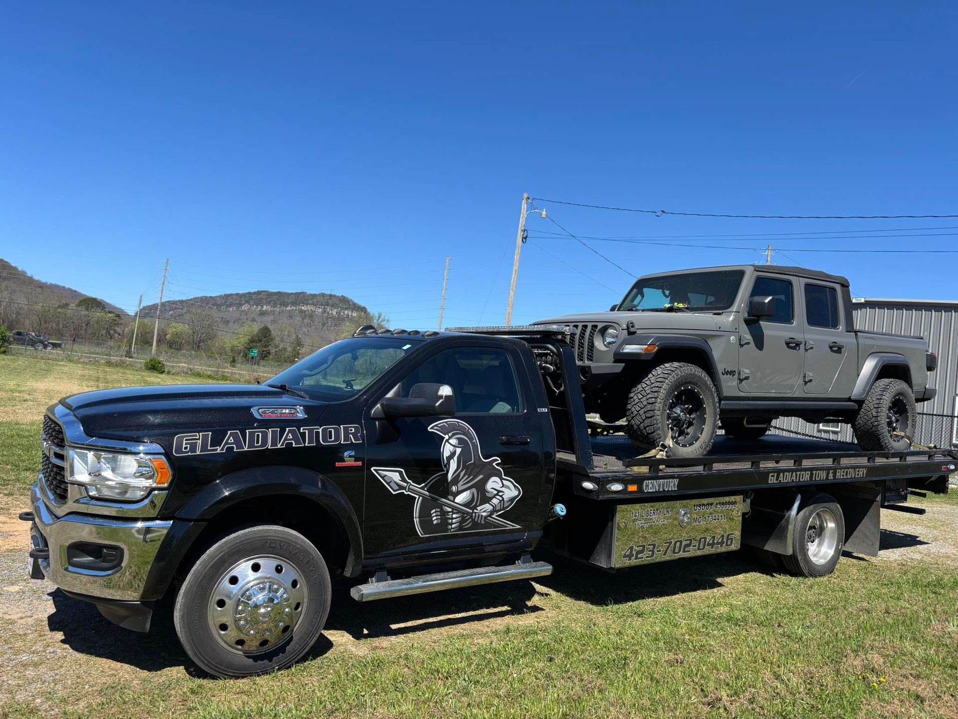 Black pickup towing a damaged gray pickup on a flatbed trailer in a sunny field.