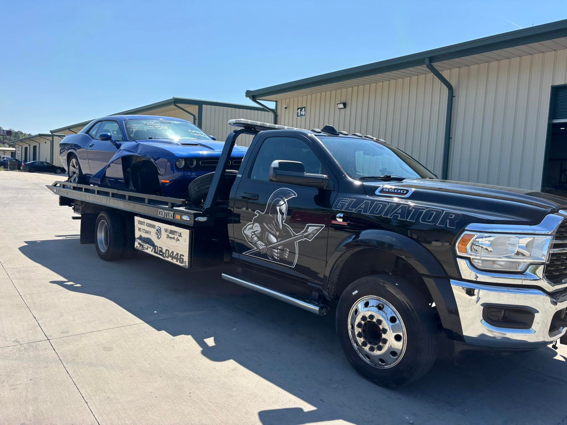 Black tow truck carrying a blue sports car on a sunny street beside a warehouse