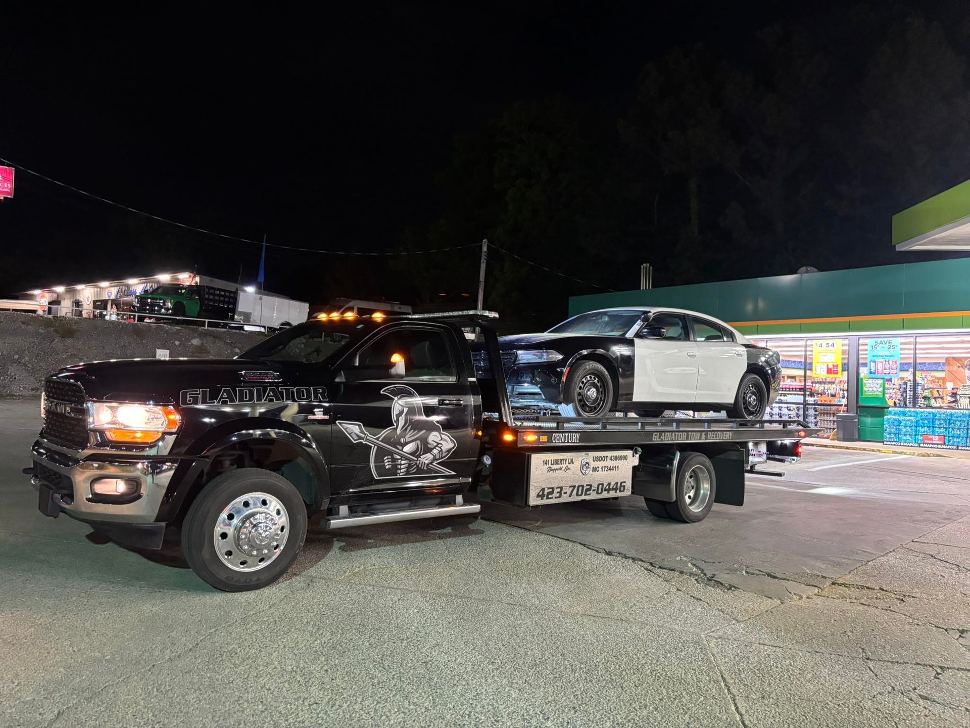 Black tow truck hauling a damaged white sedan in a lit parking lot at night