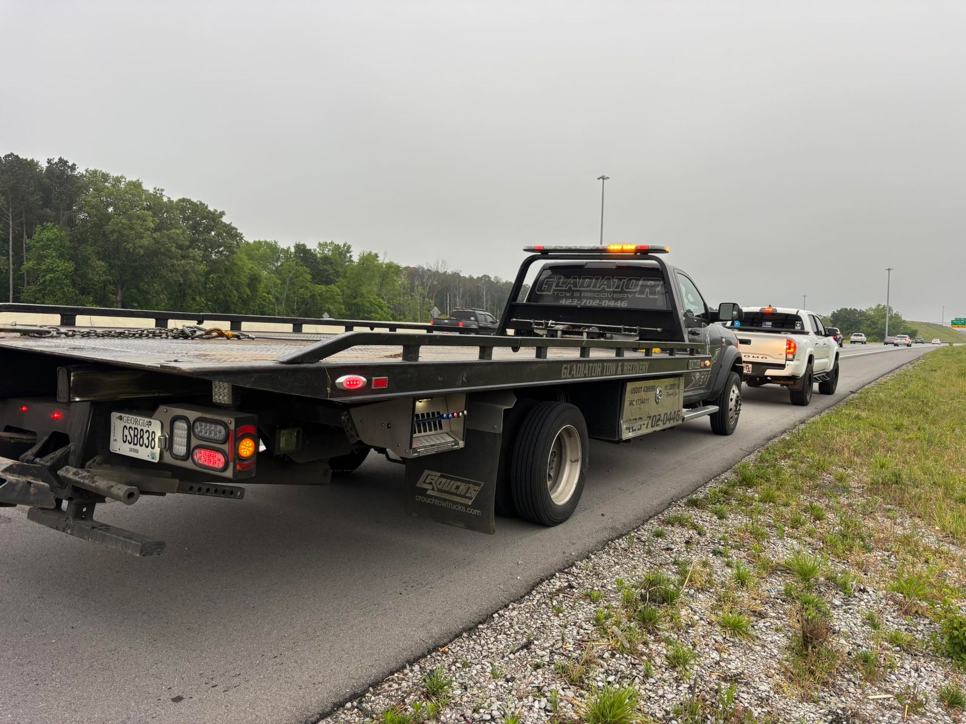 Tow truck towing a car on a wet highway shoulder, with traffic and trees in the background