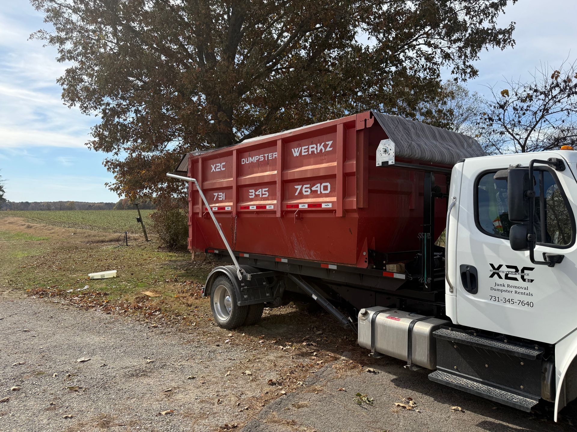 Red dumpster on a white truck parked beside a gravel road in a rural field under a cloudy sky