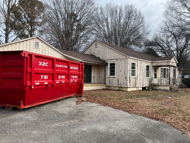 Red dumpster beside a weathered beige house on a cloudy day