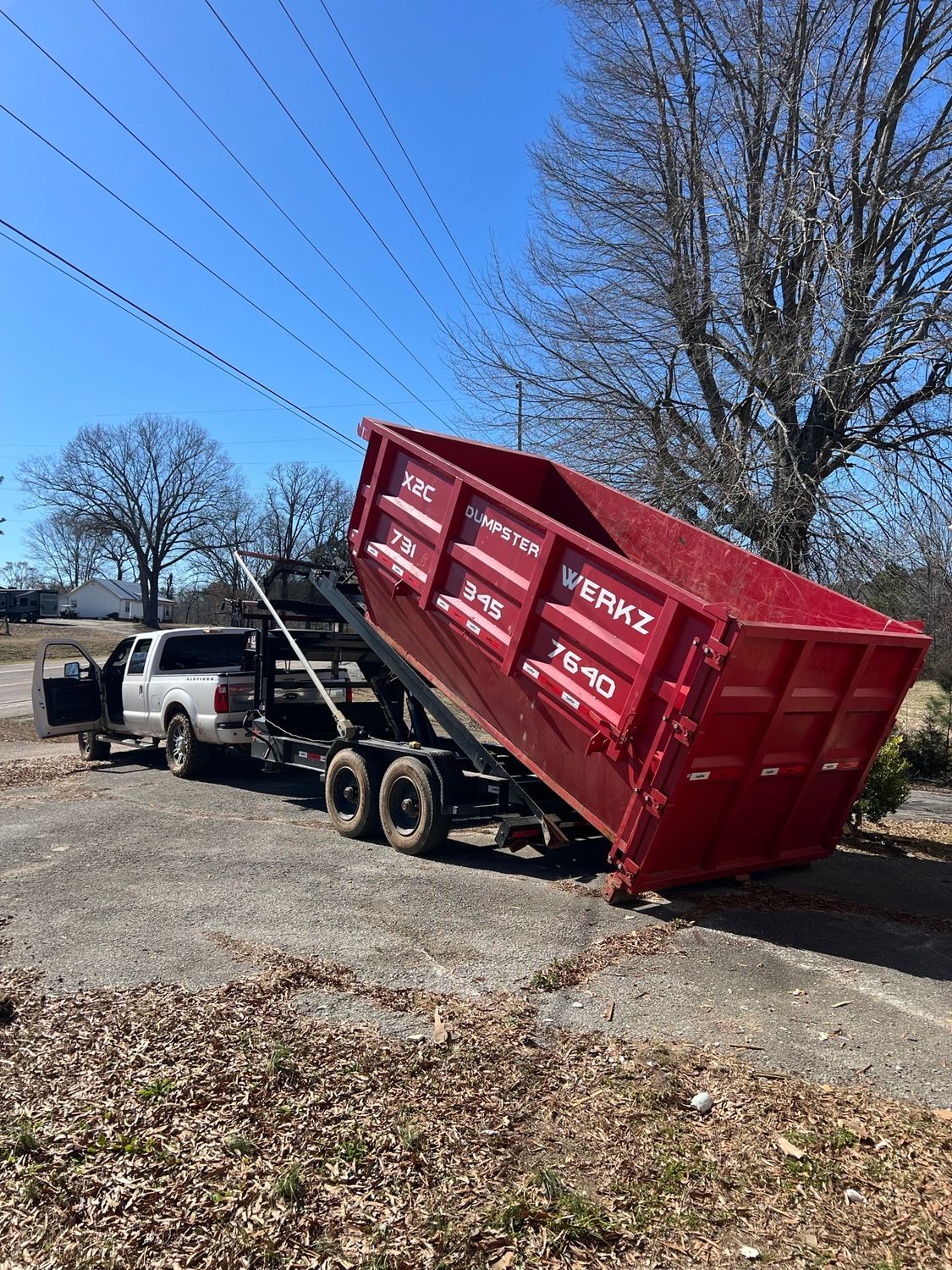 Pickup truck dumping a large red dumpster on a roadside driveway under a clear blue sky