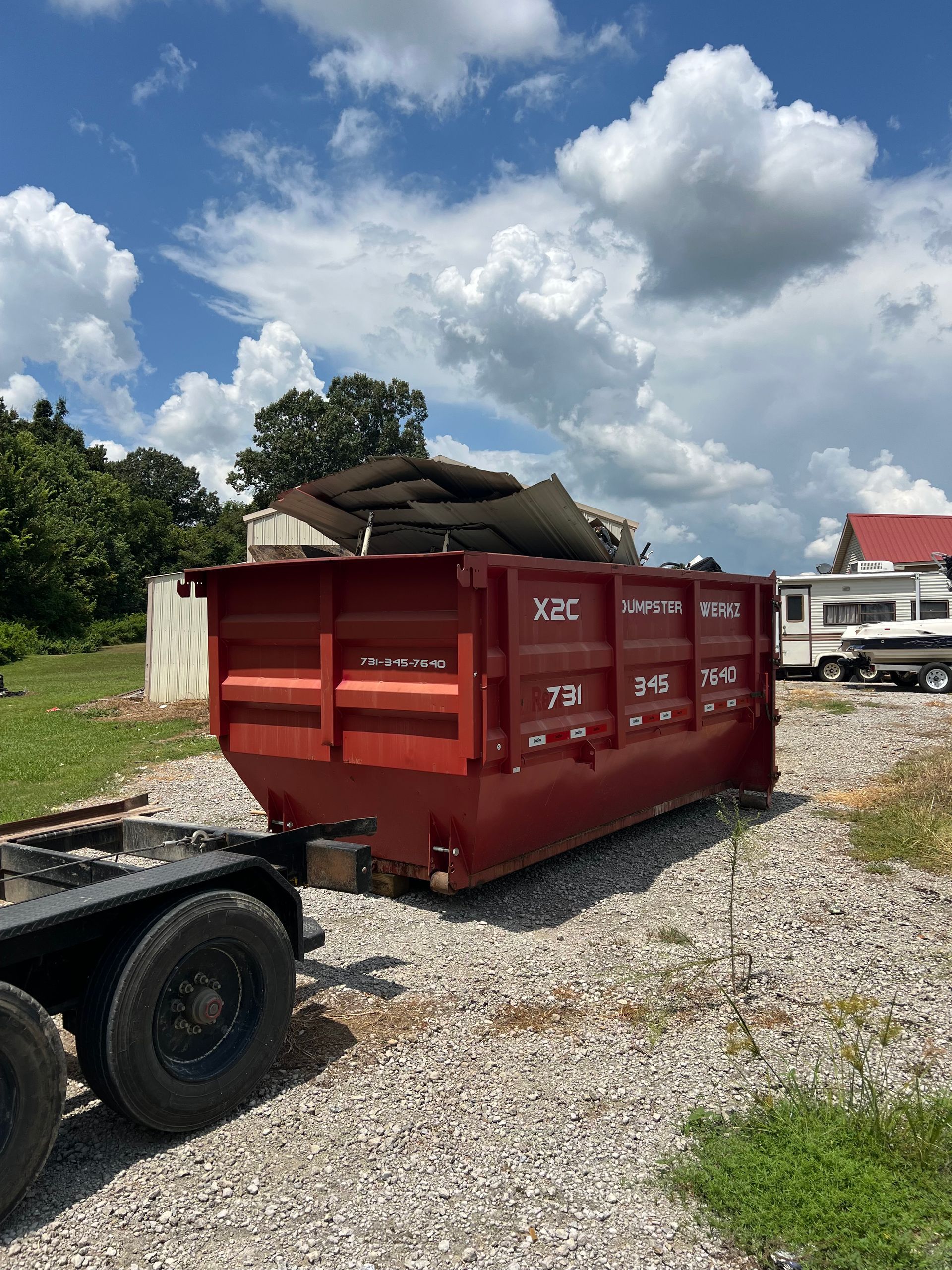 Red dumpster on gravel beside a trailer, with a house and cloudy blue sky in the background.