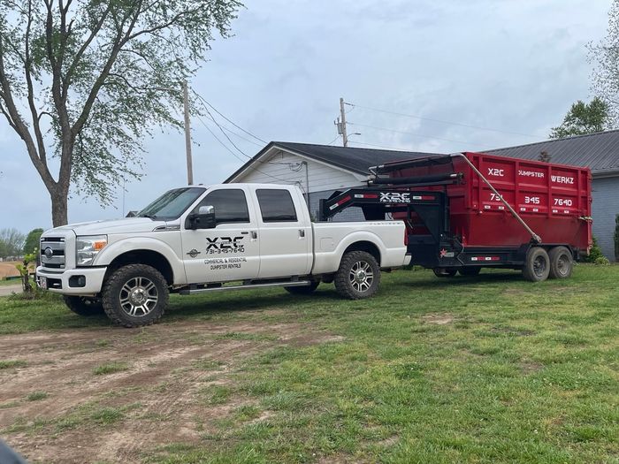 White pickup truck hauling a red dump trailer on grass beside a house under cloudy skies