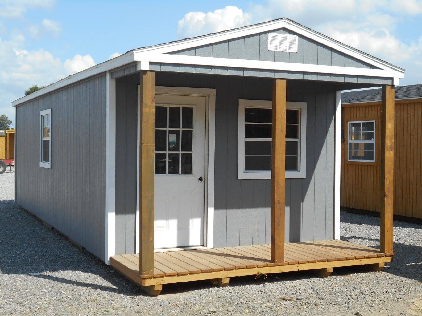 Gray shed with white trim, porch, and door; set outdoors on a gravel lot.