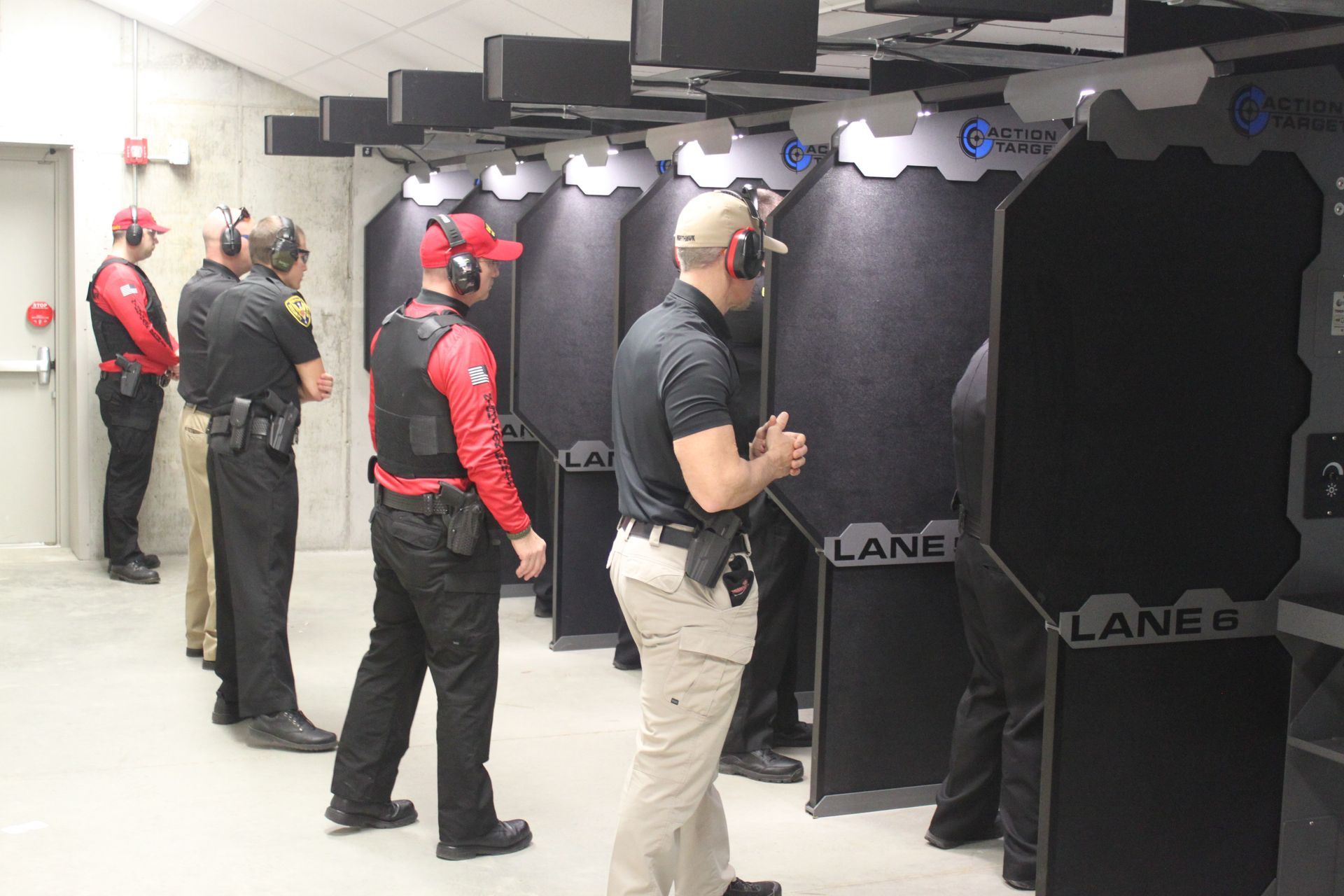 People practicing shooting at an indoor gun range. They wear hats and tactical gear, standing by shooting lanes.