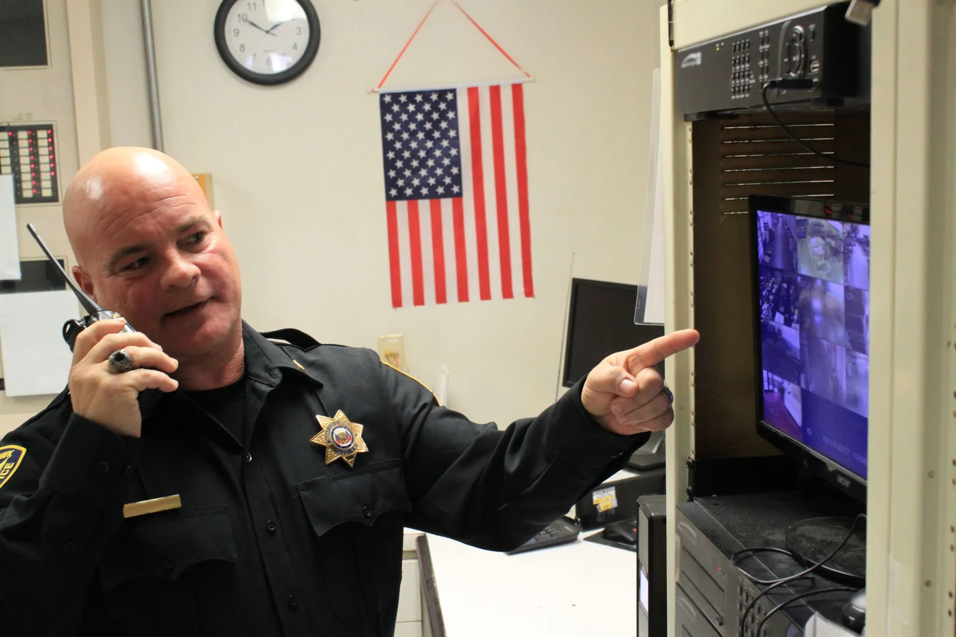 Man in sheriff’s uniform talking on phone and pointing at a TV in an office with an American flag.
