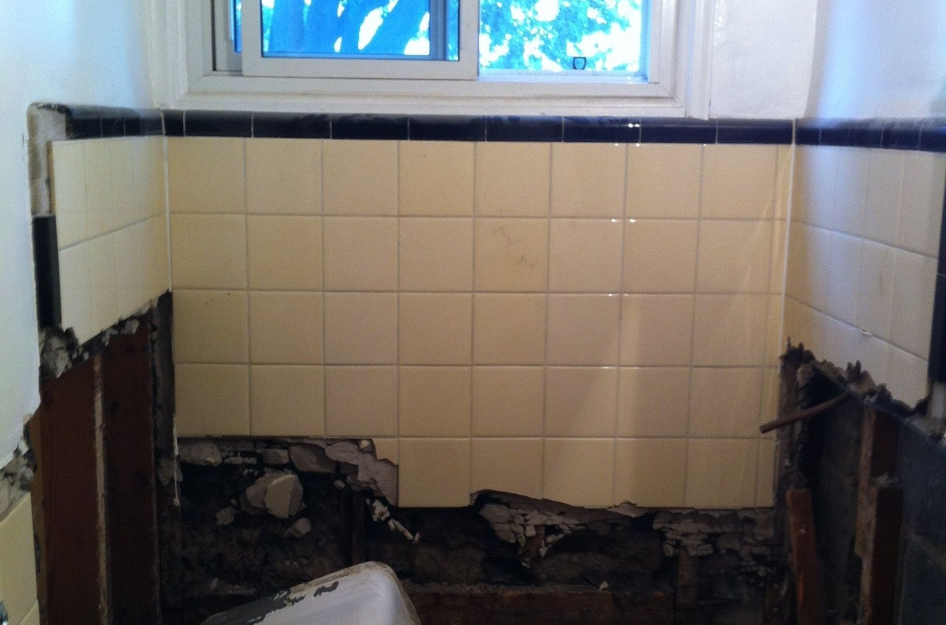 A bathroom wall undergoing renovation with partially demolished plaster around yellow tiles and a dark tiled border.