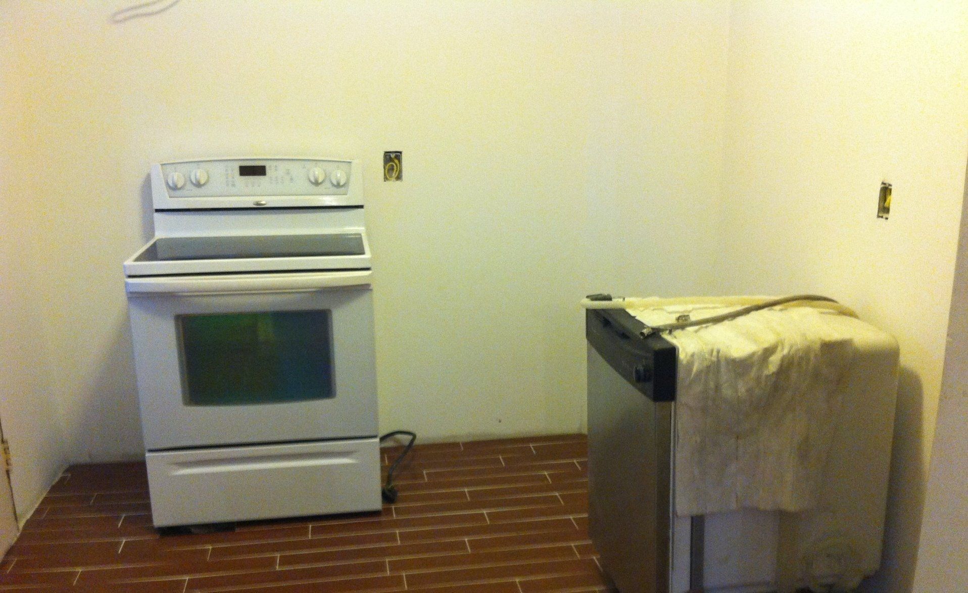 A white electric stove stands beside a stainless steel dishwasher covered with a cloth against a plain, unfurnished wall.