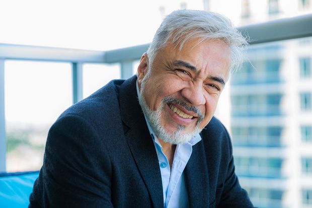 A smiling person with gray hair and a beard wearing a dark blazer over a light blue shirt on a balcony.