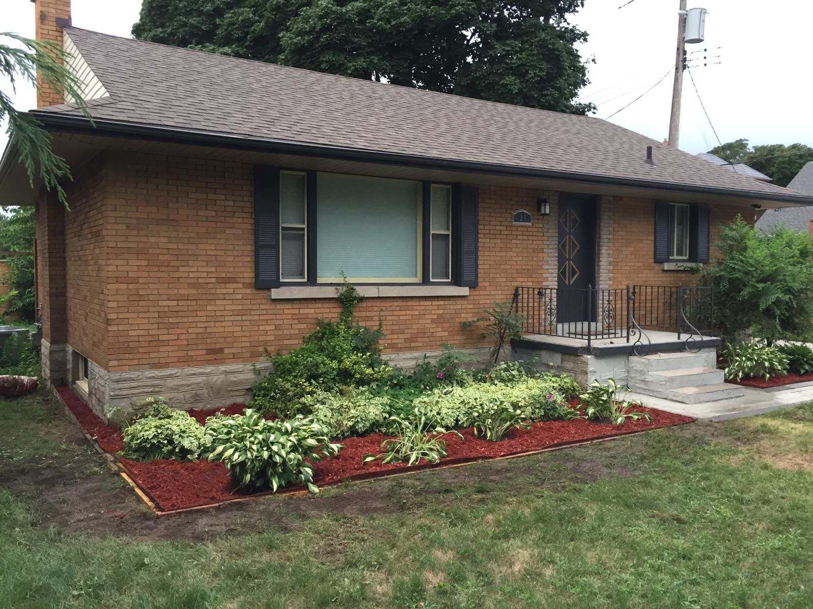 A one-story brick house with a brown shingled roof, front porch, and a mulched garden bed with plants in the foreground.