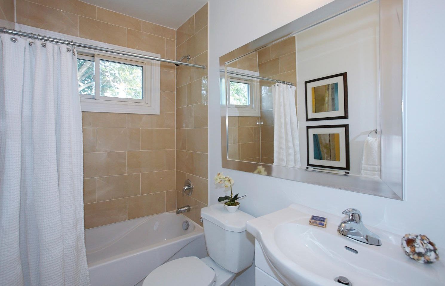 A bright, modern bathroom featuring a white porcelain sink, toilet, and a bathtub with a white curtain and beige tile.