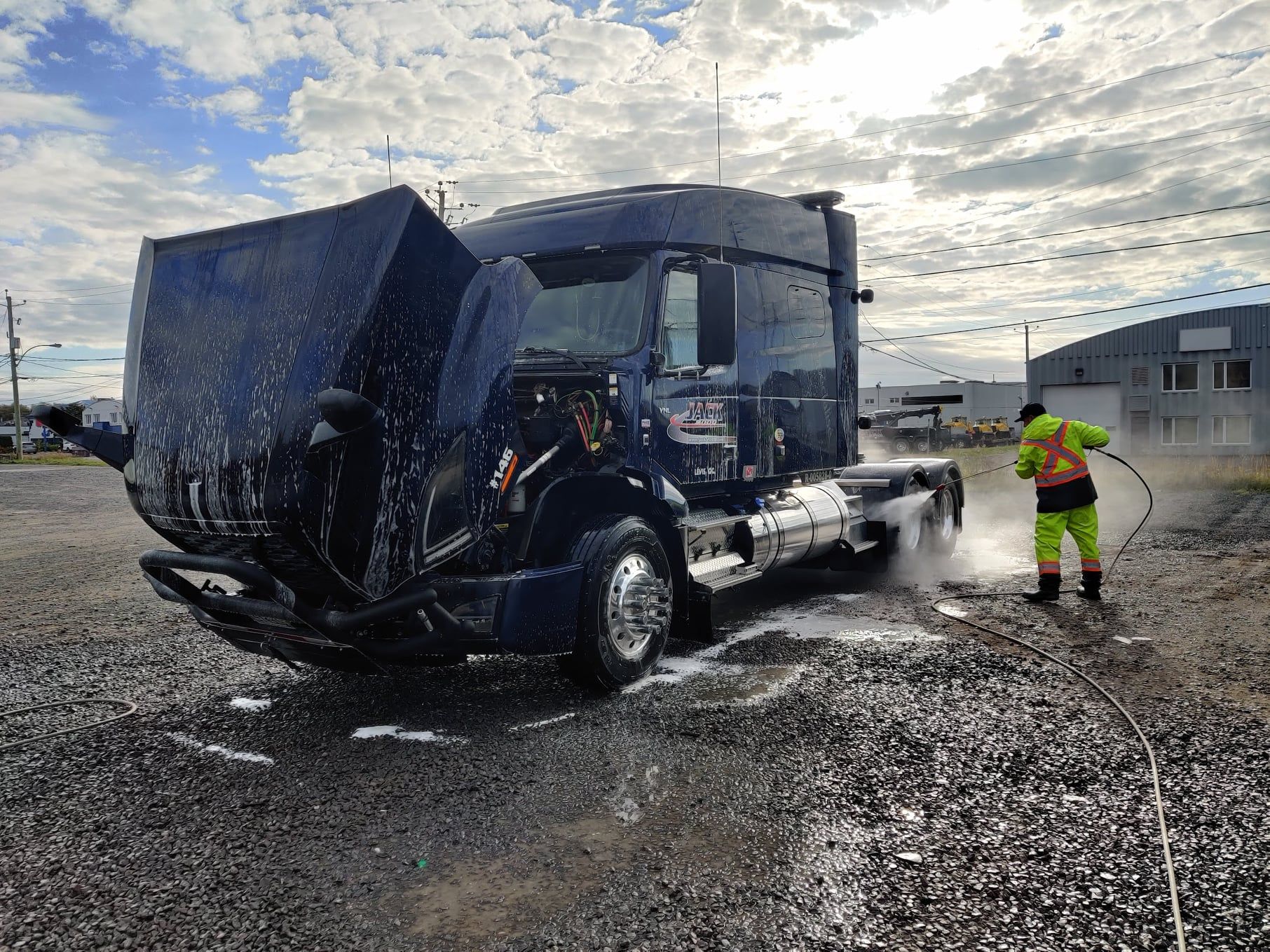 Un homme lave un camion avec un tuyau dans un parking en gravier.