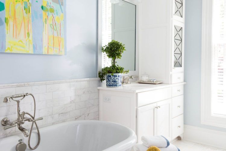 Bright white bathroom with a bathtub, wall art, and a potted plant on a cabinet by a sunlit window.