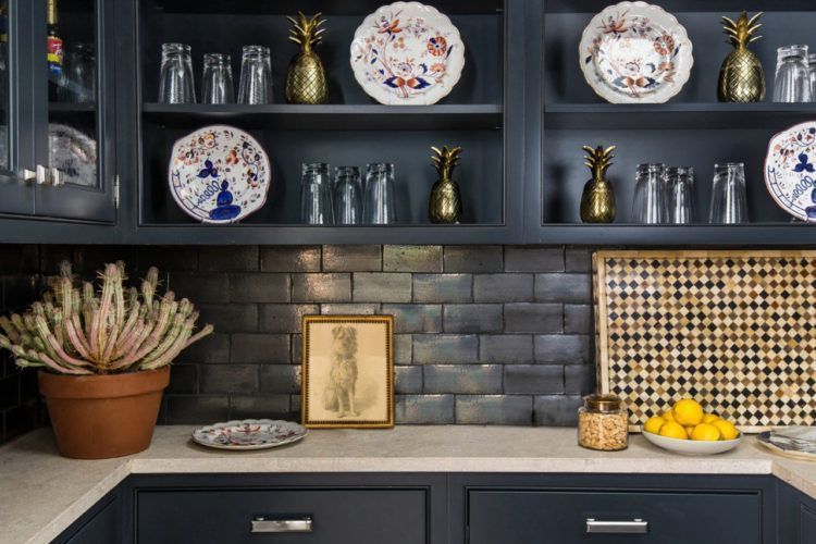 Dark kitchen shelves with decorative plates, glassware, pineapples, a potted plant, and fruit on the counter