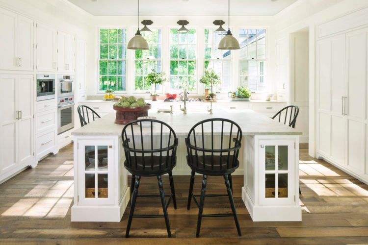 Bright white kitchen with large island, black chairs, pendant lights, and sunlight through windows