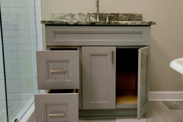 Gray bathroom vanity with granite top, left drawers open and cabinet doors ajar, beside a tiled shower.
