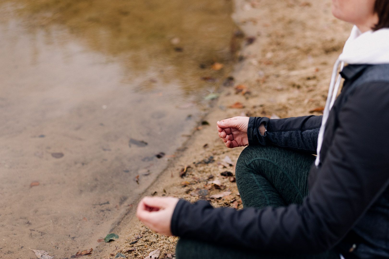 photo showing a ladies hands resting on her knees, sitting on the ground by a lake