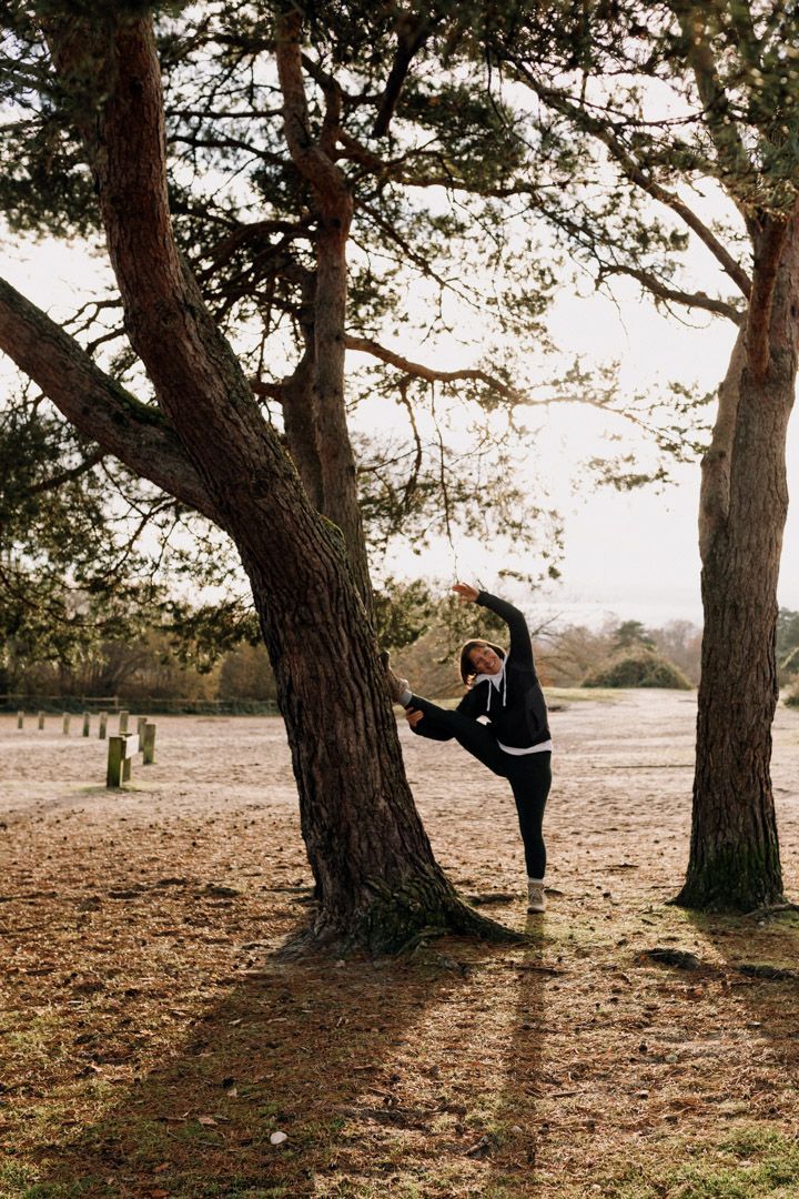 photo of a lady doing a stretch with her leg lifted high against a tree standing on sand