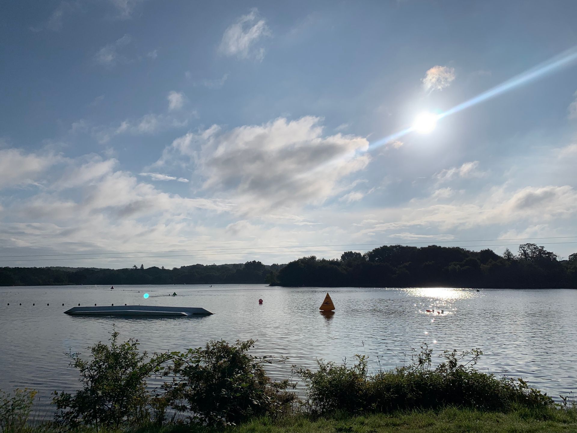 photo of a lake, with greenery in the foreground and background. The sky is blue with the sun shining from behind small clouds
