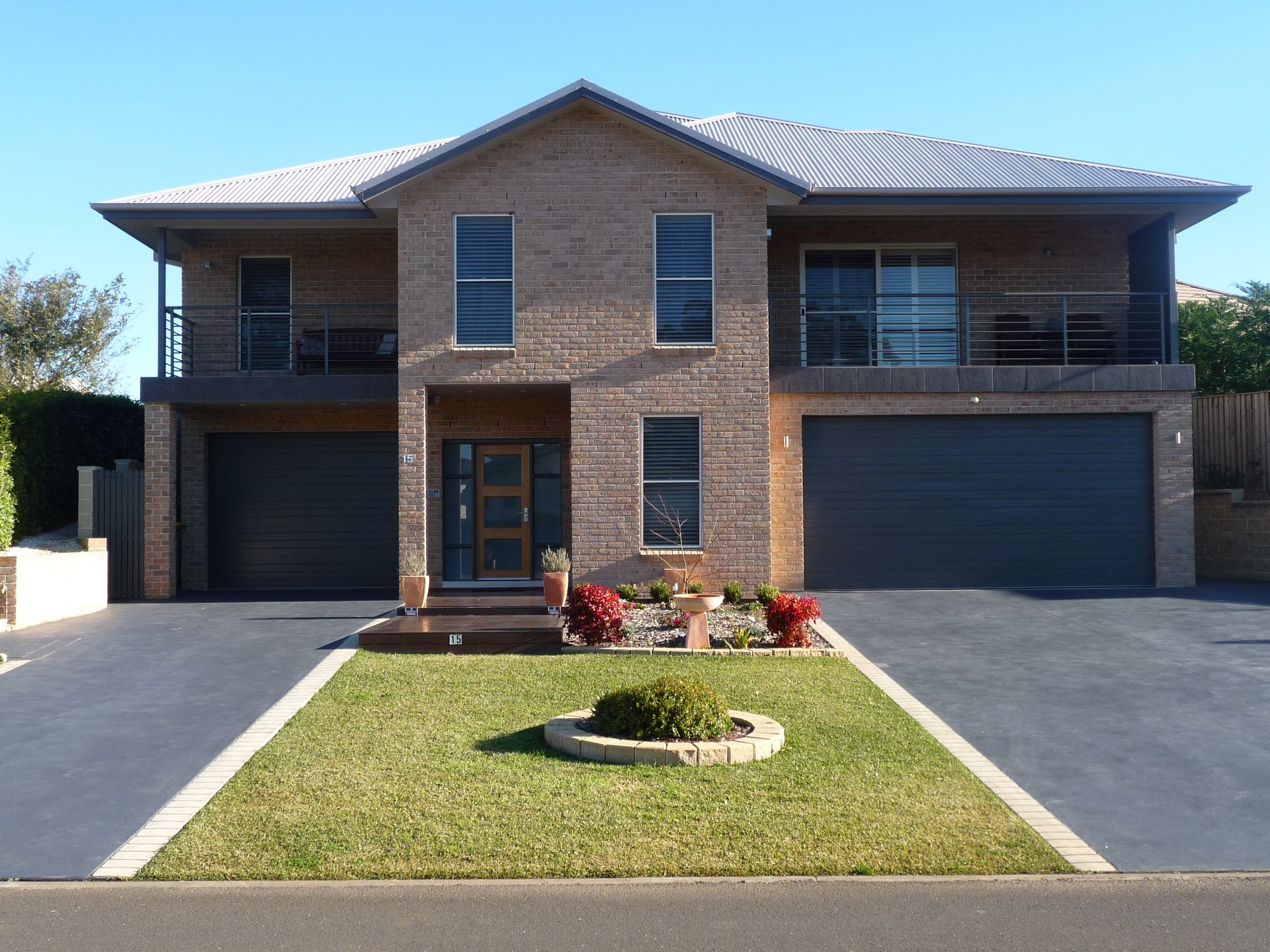 Two-story brick house with two garages, balconies, and a small front garden, blue sky.
