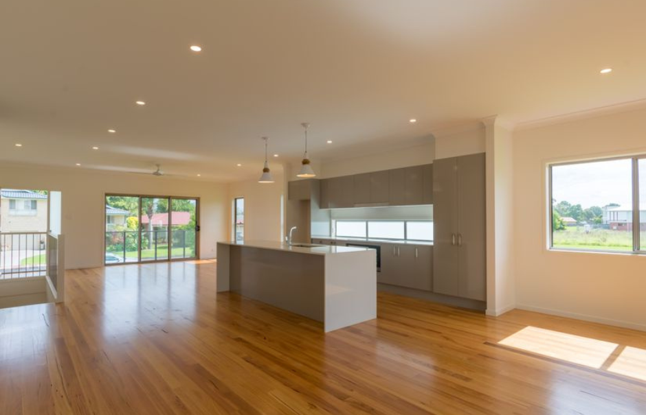Spacious empty modern kitchen and living area with hardwood floors, white island, and large windows with sunlight.