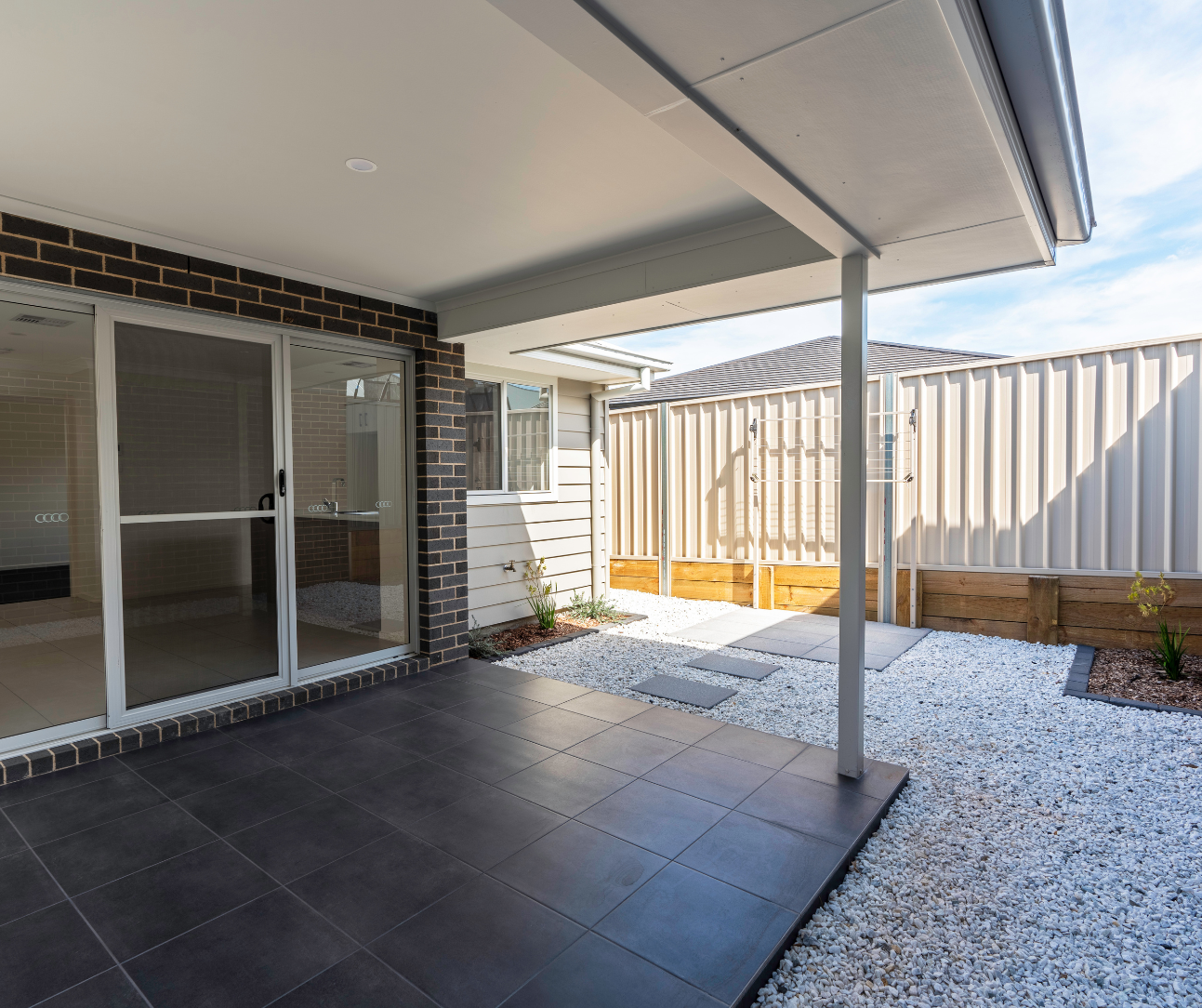 Covered patio with sliding glass door, dark tiled floor, and gravel yard beside a corrugated fence. Covered patio with sliding glass door, dark tiled floor, and gravel yard beside a corrugated fence.