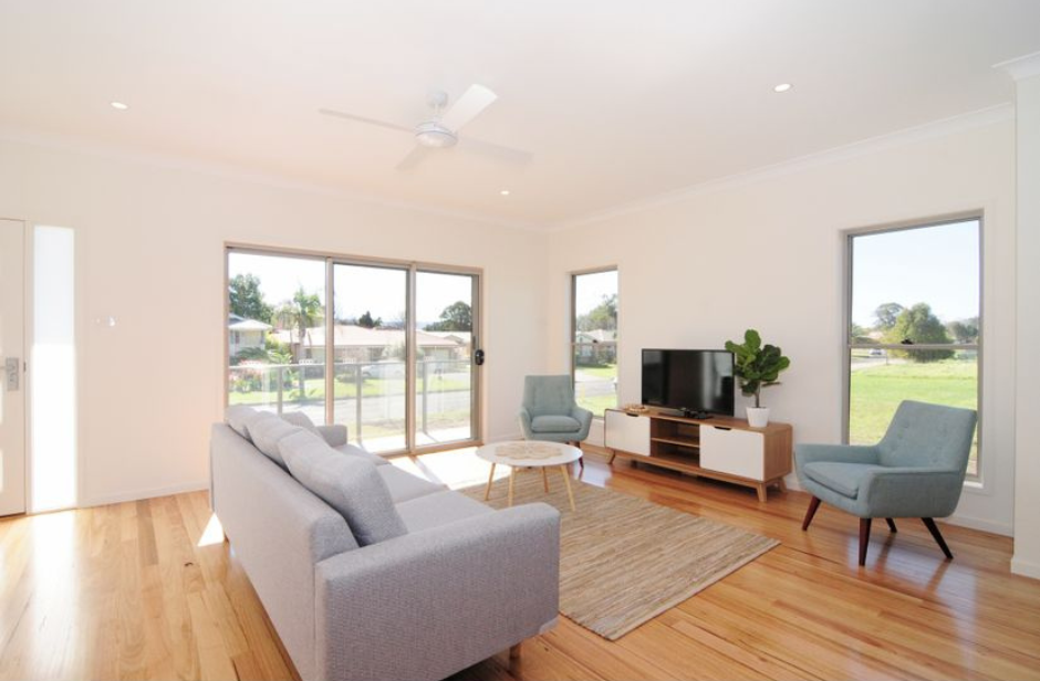 Bright modern living room with light wood floors, gray sofa, blue chair, TV, and large windows.