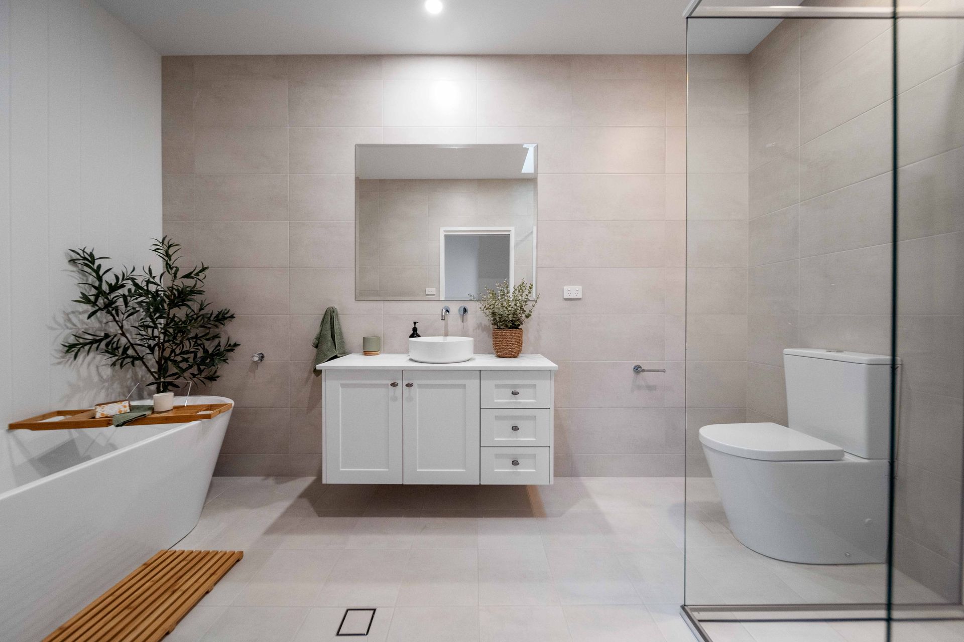 Modern bathroom with white vanity, toilet, and bathtub. Glass shower, light beige walls, and wooden accents.
