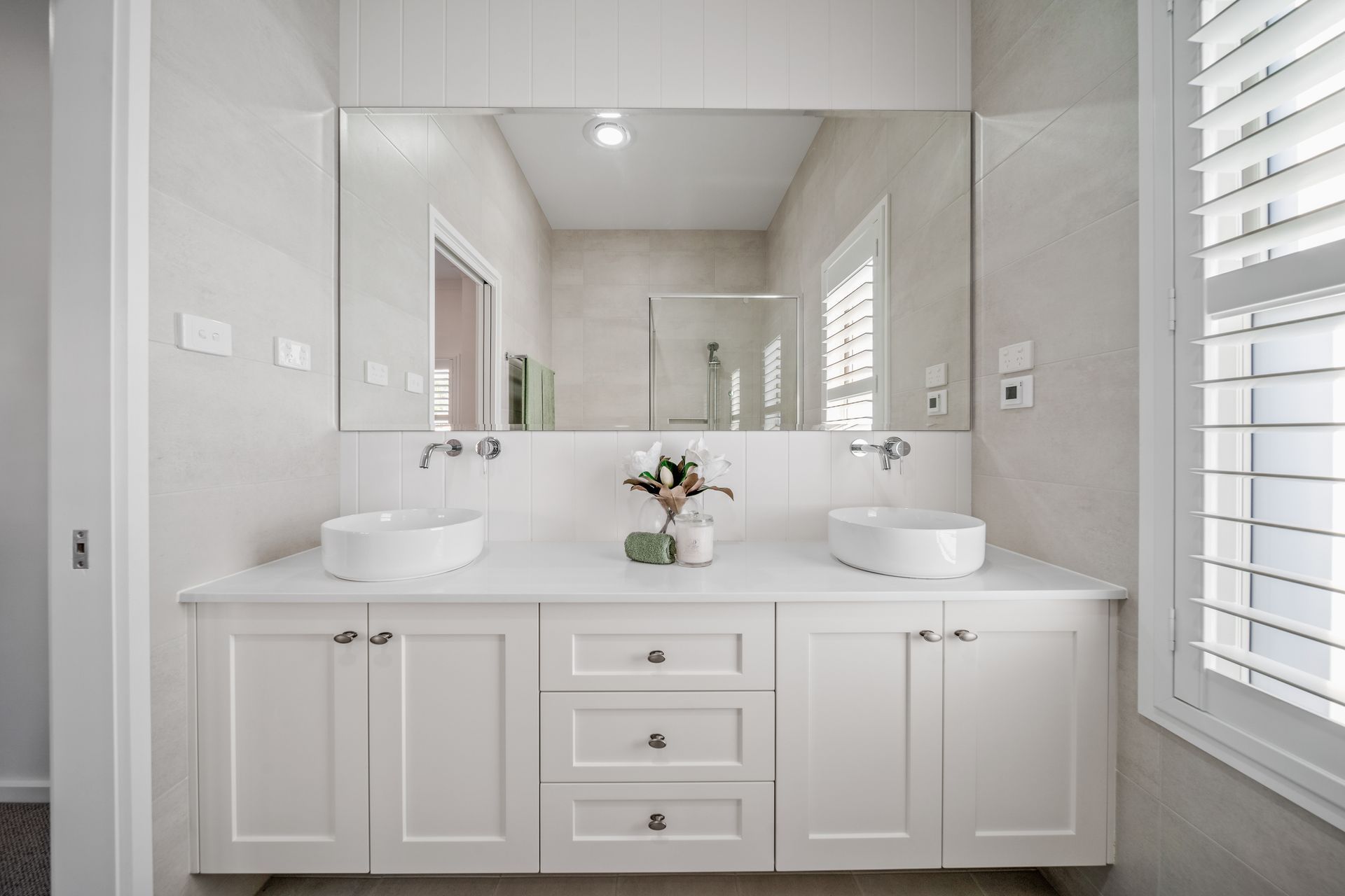 White bathroom with dual sinks, large mirror, and shuttered window.