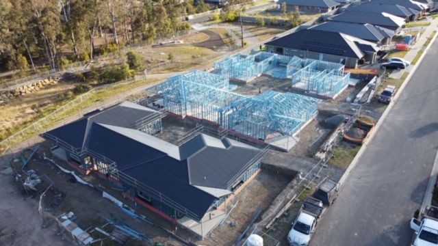 Houses under construction with exposed blue metal frames, near completed structures, and a paved road.