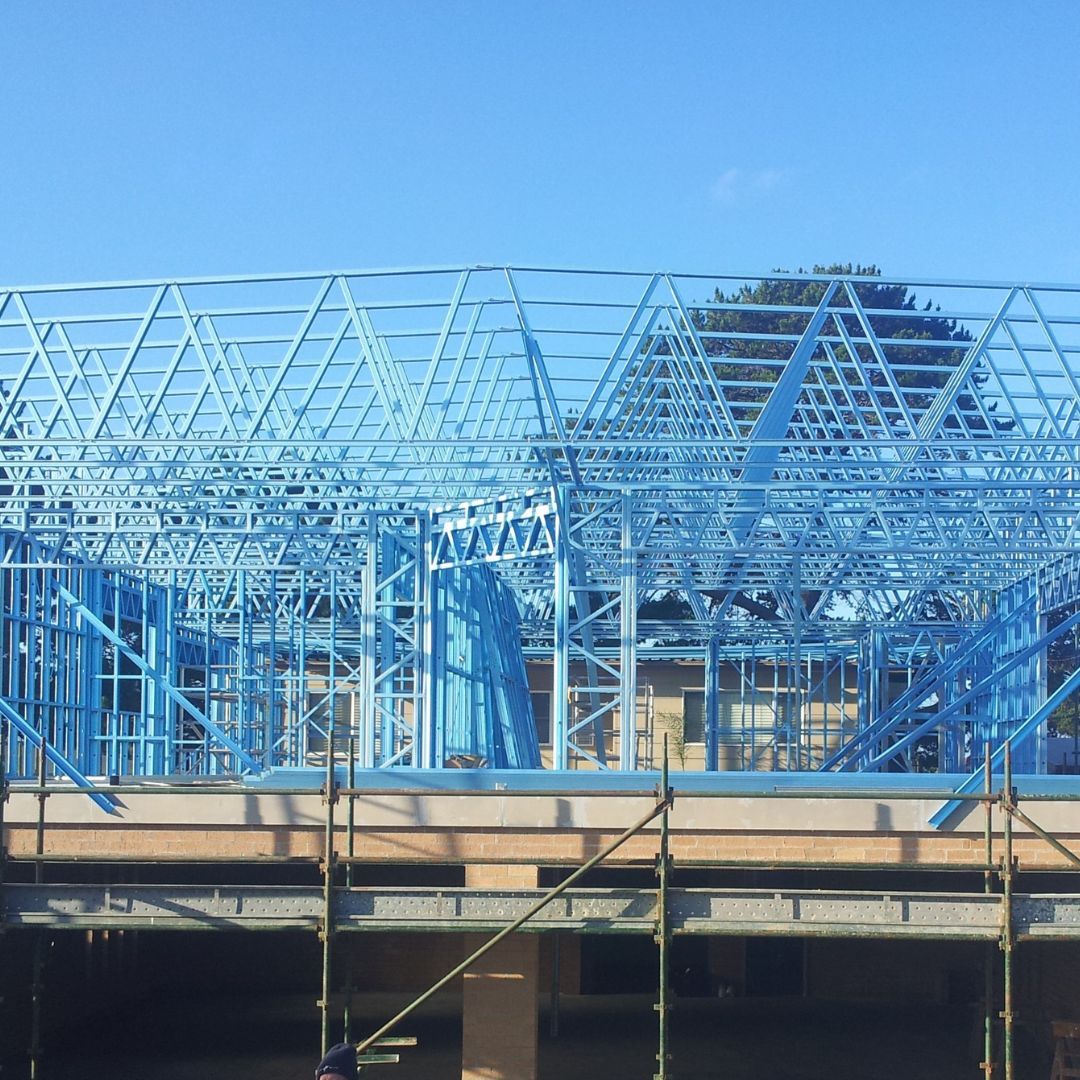 Blue steel frame of a house under construction; roof trusses and scaffolding visible, clear blue sky.