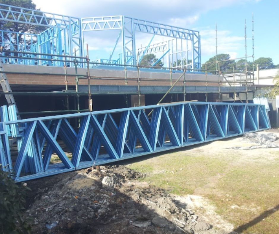 Blue steel frame construction, with a partially built building and a truss lying on the ground. Outdoors, sunny day.