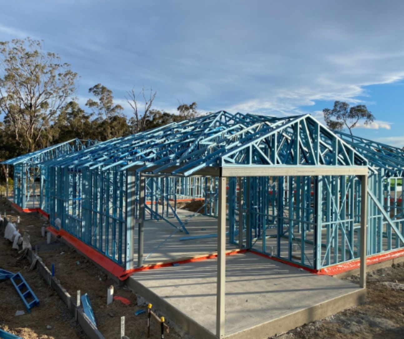 Blue steel frame of a house under construction on a concrete foundation, with a blue sky in the background.