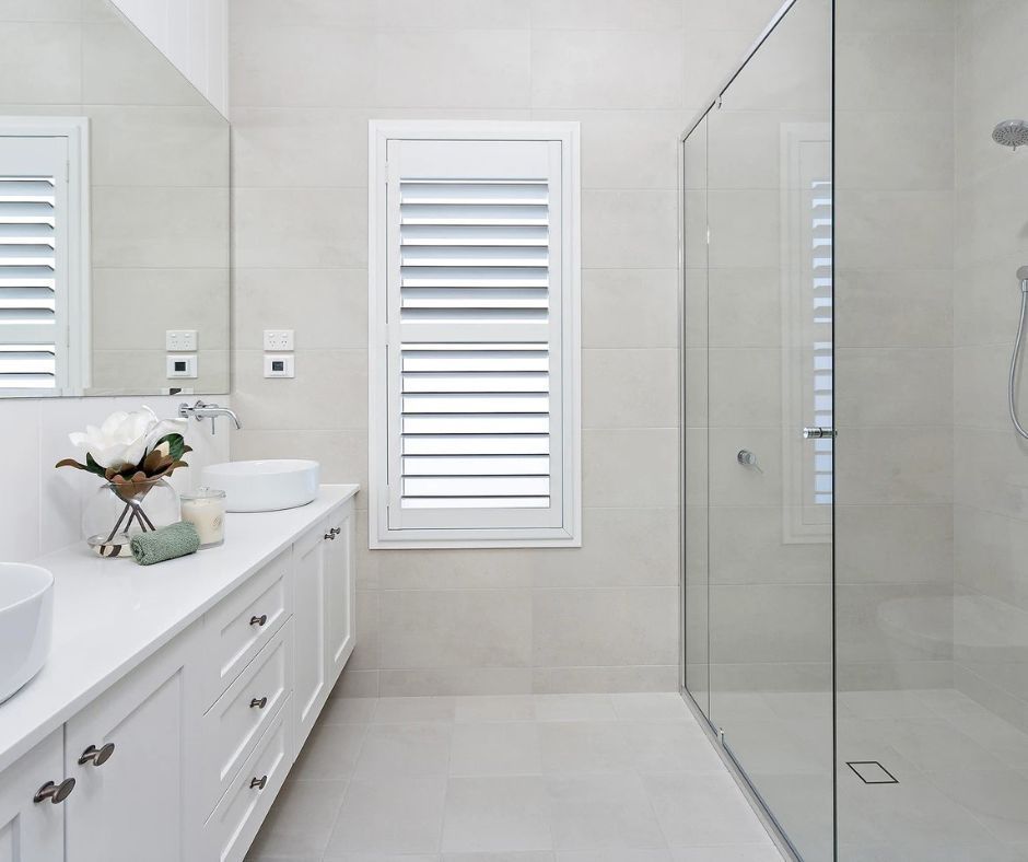 Modern white bathroom with vanity, shower, and window with shutters.