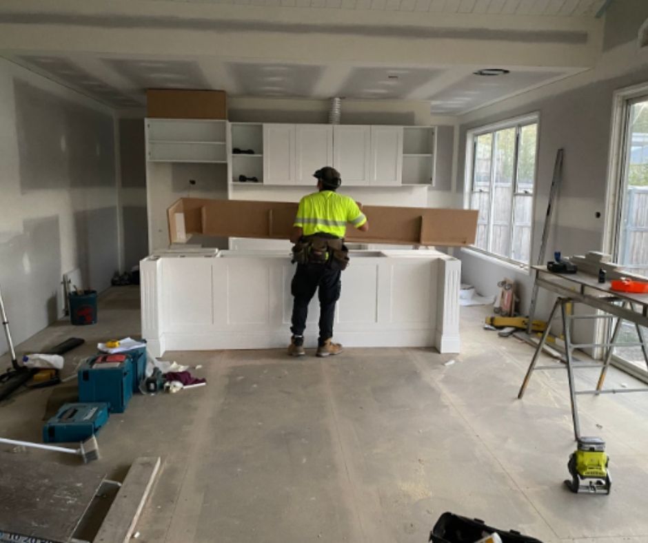 A person in a yellow vest works on a kitchen island during construction; white cabinets and drywall are visible.