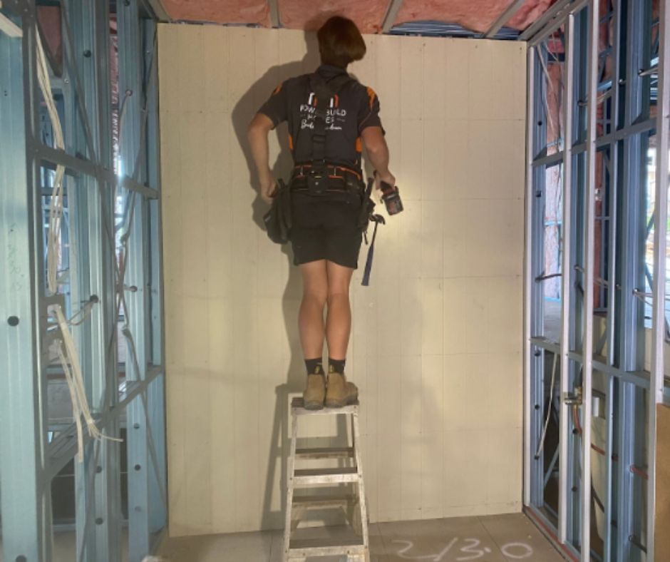 Construction worker standing on a step stool, working on drywall inside a building frame.