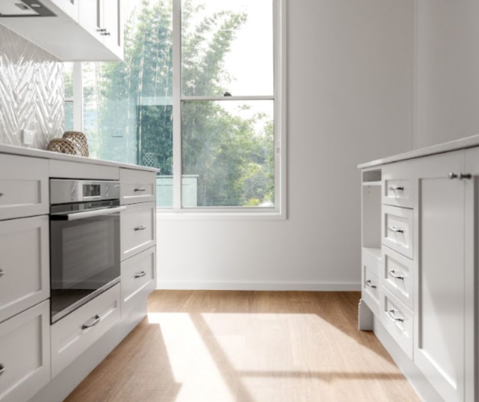 White kitchen with built-in oven and cabinets, next to a window with sunlight and wood floors.